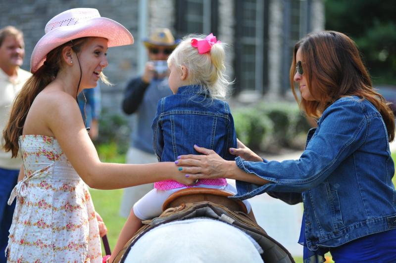 small pony rider being secured in saddle by pony walker and by her mother